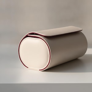 Può includere: Un astuccio cilindrico con esterno liscio color crema e rivestimento interno bordeaux scuro. L'astuccio presenta una chiusura a patta che si estende sulla parte superiore, con cuciture visibili lungo i bordi del pannello laterale circolare.