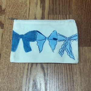 Könnte beinhalten: Eine rechteckige, cremeweiße Canvas-Tasche mit Reißverschluss, die auf der Vorderseite eine horizontale Reihe aus drei verschiedenen blauen Schleifen zeigt. Die erste Schleife ist dunkelblau strukturiert, die zweite hellblau und die dritte ein hellblaues Band.