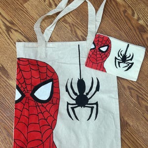 May include: A beige canvas tote bag and a matching rectangular zippered pouch. Both items feature a red and black spider-web mask design on the left side and a black spider graphic hanging from a vertical line on the right side of the fabric.