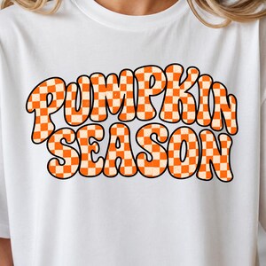 May include: A white short-sleeved t-shirt featuring the text PUMPKIN SEASON in a bold, wavy, retro-style font. The letters are filled with an orange and white checkered pattern and outlined in black, centered on the chest of the garment.