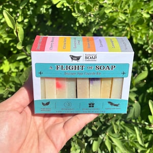 Puede incluir: Una caja rectangular etiquetada como A FLIGHT OF SOAP que contiene ocho pastillas de jabón pequeñas. El empaque presenta una franja superior codificada por colores con nombres de aromas y una sección inferior con iconos. Cada pastilla pesa 28 g.