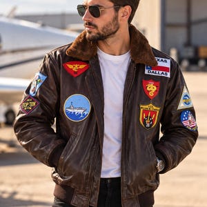 Puede incluir: Una chaqueta bomber de cuero marrón con cuello de piel sintética marrón, una camiseta blanca y vaqueros negros. La chaqueta presenta varios parches bordados, incluyendo una bandera de Texas Dallas, un emblema del U.S.S. William M. Standley y un parche Top Ten.