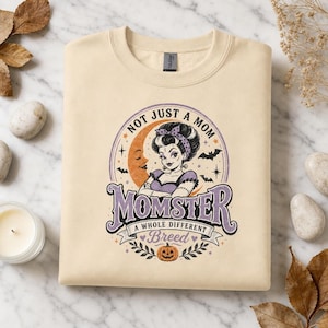 Könnte beinhalten: Ein beigefarbenes Sweatshirt mit Rundhalsausschnitt und einem Grafikdruck einer Frau mit violettem Stirnband, einer Mondsichel, Fledermäusen und einem Kürbis. Der Text lautet: NOT JUST A MOM, MOMSTER, A WHOLE DIFFERENT BREED.