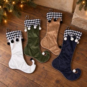 May include: Four velvet Christmas stockings in white, olive green, tan, and navy blue. Each stocking features a black and white checkered cuff with scalloped edges and small white or black pom-poms. The stockings have a curled toe design.