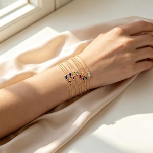 May include: A set of delicate gold chain bracelets, each featuring a single rectangular baguette-cut gemstone in various colors including blue, red, green, yellow, and clear, arranged in a stack on a wrist.