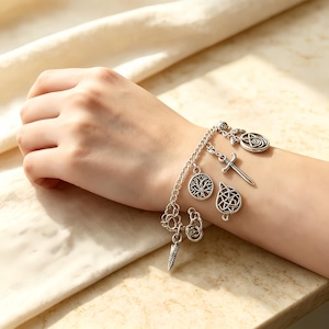May include: A silver-toned charm bracelet featuring multiple dangling pendants, including a tree of life, a sword, a teardrop-shaped knot design, and a circular spiral emblem, all attached to a delicate metal chain link band.