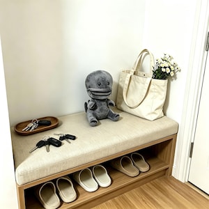 Peut inclure: Un banc d'entrée en bois clair avec une assise beige rembourrée, comprenant une peluche grise en forme de créature avec une bouche à fermeture éclair, un sac fourre-tout en toile crème avec des fleurs blanches, un plateau en bois et des chaussons.