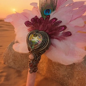 May include: A handheld fan featuring light pink feathers and a single peacock feather. The handle is adorned with a heart-shaped glass centerpiece, intricate bronze-toned metal scrollwork, and embedded pink and dark red oval-shaped stones.