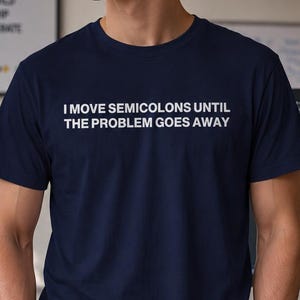 Könnte beinhalten: Ein dunkelblaues Kurzarm-T-Shirt mit dem weißen Text I MOVE SEMICOLONS UNTIL THE PROBLEM GOES AWAY auf der Brust. Das Kleidungsstück hat einen klassischen Rundhalsausschnitt und einen geraden Saum, was für ein schlichtes und sauberes Aussehen sorgt.