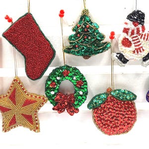 Peut inclure: Une collection d'ornements de fête perlés comprenant des sucres d'orge, une chaussette rouge, un sapin de Noël vert, un bonhomme de neige, une cloche, une étoile dorée, une couronne, une pomme rouge et une grappe de raisins violets, ornés de sequins.