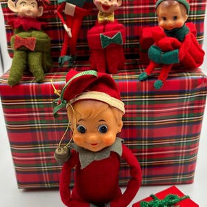 May include: Five elf figurines with painted faces, pointed ears, and red and green clothing. Four elves are positioned on a red and green plaid gift box, while one sits in front with a small gold bell attached to its hat. A small red gift box sits nearby.