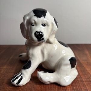 May include: A ceramic dog figurine featuring a white base with black spots and a glossy crackle glaze finish. The puppy is depicted in a seated position with floppy ears, a black nose, and dark eyes, showcasing a detailed, smooth surface texture.