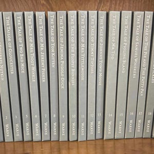May include: A collection of twenty-three gray hardcover books titled The Tale of Peter Rabbit and other stories by Beatrix Potter, published by Warne, arranged in a row between two ceramic rabbit figurines on a wooden shelf.