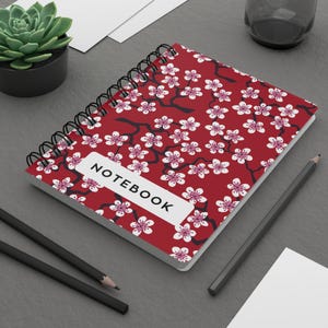 Puede incluir: Un cuaderno de espiral con una cubierta de color rojo intenso que presenta un patrón repetitivo de flores de cerezo blancas y rosas sobre ramas oscuras. Una etiqueta rectangular blanca en la portada muestra la palabra NOTEBOOK en letras negras.
