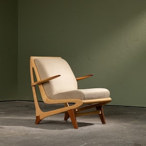 Op de afbeelding: Een fauteuil met een lichtbeige gestoffeerde zitting en rugleuning, ondersteund door een licht houten frame met opvallend gebogen, hoekige poten en slanke, puntige armleuningen die naar buiten uitsteken vanaf de rugstructuur.