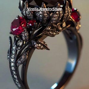 May include: A black metal ring featuring a large dark centre stone, vibrant red round side stones, and small clear pavé accents shaped like leaves. The text VirellaJewelryShop is visible on the band. The design incorporates intricate metalwork.