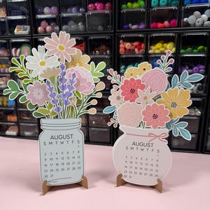 May include: Two decorative paper calendars for August, each featuring a colorful floral arrangement. The left calendar is shaped like a mason jar, while the right one is a rounded vase with a small bow. Both display the August calendar grid.