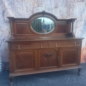 Könnte beinhalten: Ein braunes Holzsideboard mit einem zentralen ovalen Spiegel, der in eine dekorative Rückwand eingelassen ist. Der Unterbau umfasst zwei Schubladen mit kleinen Messinggriffen und drei untere Schranktüren, wobei die mittlere Tür ein geschnitztes Blumenmotiv aufweist.