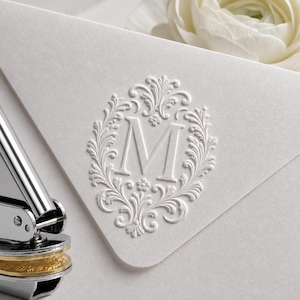 May include: A white paper envelope featuring a central embossed letter M surrounded by an intricate floral scrollwork design. A silver metal hand embosser tool is positioned at the bottom left corner, showing its metallic construction and circular base.