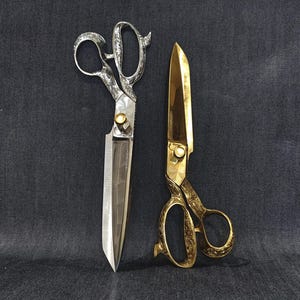 May include: A pair of silver-toned metal shears with an ornate handle and a gold-toned screw, positioned next to a pair of gold-toned metal shears featuring an intricate floral-style embossed pattern on the handles and blades.