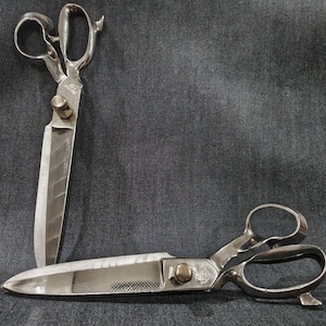 Könnte beinhalten: Zwei Paar silberfarbene Schneiderscheren aus Metall mit großen, ergonomischen Griffen und einer zentralen Drehschraube. Eine Schere steht vertikal, während die andere horizontal liegt und scharfe, polierte Klingen sowie eine texturierte Grifffläche zeigt.