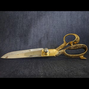 May include: A pair of gold-colored metal scissors featuring intricate floral engravings on the handles and a polished finish. The blades are metallic silver with a sharp point, connected by a central silver-toned screw mechanism.