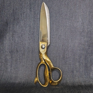 May include: A pair of gold-coloured metal scissors featuring a polished finish on the handles and a silver-toned blade. The scissors have a central screw mechanism connecting the two blades and two asymmetrical finger loops for manual operation.
