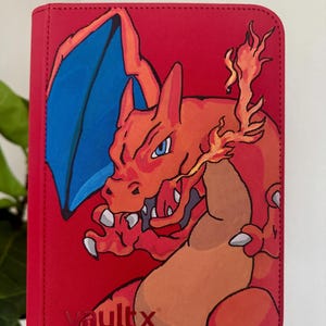 May include: A red trading card binder featuring a large, orange, dragon-like creature with blue wings and flames coming from its mouth. The cover has a textured surface and the word vaultx printed in the bottom left corner in a dark red font.