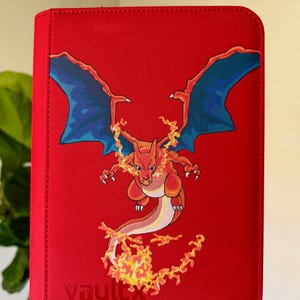 May include: A bright red zippered binder featuring a graphic of a fire-breathing orange dragon with large blue wings. The dragon is surrounded by flames, and the word vaultx is embossed in the bottom left corner of the textured red cover.