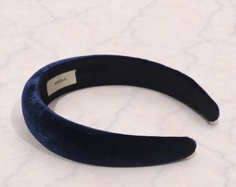 Puede incluir: Una diadema gruesa y acolchada de tela de terciopelo azul marino intenso. El interior de la diadema presenta una pequeña etiqueta de tela blanca con el texto 100% SILK impreso en negro, indicando la composición del material del accesorio.