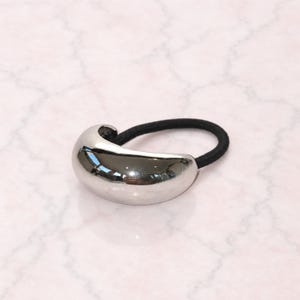 May include: A hair tie featuring a smooth, polished silver-tone metal accent piece with a curved, bean-like shape. The metal component is attached to a simple, circular black elastic band, creating a functional hair accessory.