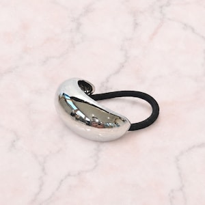 May include: A hair tie featuring a smooth, polished silver-tone metallic bead with a curved, bean-like shape attached to a simple, solid black elastic band. The reflective surface of the metal bead shows light highlights.
