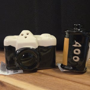 以下が含まれることがあります： ヴィンテージカメラと35mmフィルムの形をしたセラミック製の塩コショウ入れセット。カメラは白と黒で、上部に3つの穴があります。フィルムケースは黒と黄色で、垂直に白い文字で「400」とプリントされています。