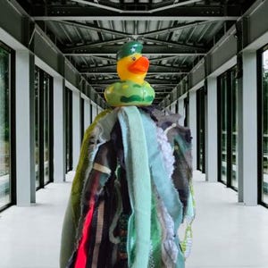 May include: A yellow rubber duck with an orange beak wearing a green camouflage beret and matching camouflage body pattern, attached to a hanging decoration made of numerous long, thin fabric strips in shades of green, black, white, and neon pink.