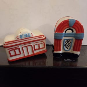 May include: A pair of ceramic salt and pepper shakers shaped like a diner and a jukebox. The diner is white with red trim and the word DINER printed in blue. The jukebox is white, red, and blue with a black base and a grid pattern on the front.
