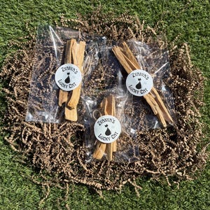 May include: Three clear plastic bags each containing several light brown wooden sticks. Each bag is sealed with a round white sticker featuring a black cat silhouette and the text Zyanya's Lucky Cat. The sticks are tied together with twine.