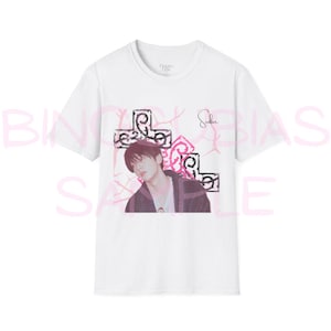 May include: A white short-sleeved t-shirt featuring a graphic print of a person with dark hair wearing a dark jacket. The design includes black decorative cross-like shapes, pink abstract patterns, and a signature script above the person.