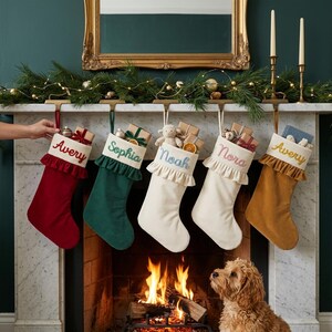 May include: Five personalized Christmas stockings hanging from a mantel, featuring the names Avery, Sophia, Noah, Nora, and Avery embroidered on the cuffs. The stockings are red, green, cream, and gold, each with a ruffled top and small gifts.
