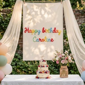 Könnte beinhalten: Ein cremefarbenes Stoffbanner, das an einem Seil hängt, zeigt den bunten, mit Garn gestickten Text Happy birthday Caroline. Darunter steht eine dreistöckige Naked-Cake-Torte mit einem C-Topper neben einer geflochtenen Vase mit rosa und weißen Blumen.