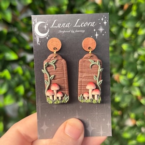 May include: A pair of rectangular dangle earrings featuring a brown wood-grain texture, green climbing vines, and small red-capped mushrooms at the base. The earrings are attached to a black display card with the text Luna Leora Inspired by fantasy.