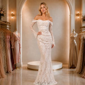 May include: A floor-length off-the-shoulder wedding dress featuring long lace sleeves and a fitted bodice. The gown is crafted from white floral lace layered over a nude-toned fabric, creating a form-fitting silhouette that extends to the floor.