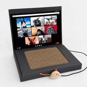 Puede incluir: Un rascador para gatos con forma de portátil negro que muestra una videollamada de ocho gatos en la pantalla. La base incluye una superficie de cartón marrón texturizado y un pequeño ratón de peluche beige unido por un cordón negro.