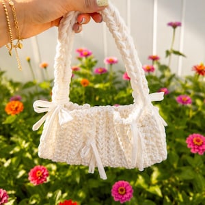 May include: A cream-colored crocheted handbag featuring a thick braided handle and decorative side ribbons. The bag has a textured, ribbed stitch pattern throughout its rectangular body, with small fabric ties cinched at the top corners.