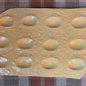 Peut inclure: Un plateau à œufs rectangulaire en céramique jaune pâle avec une finition brillante et un motif en relief de volutes tourbillonnantes sur toute la surface. Le plateau comporte douze empreintes ovales lisses disposées en trois rangées de quatre.