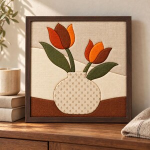 May include: A square framed fabric wall art piece featuring a textile applique of two tulips in shades of orange and brown with green leaves. The flowers are placed in a cream-colored vase with a small brown pattern, set against a beige background.