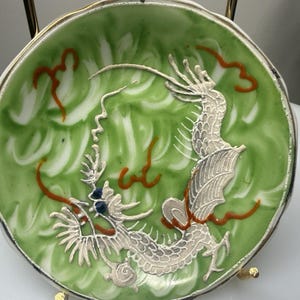 May include: A round ceramic dish featuring a raised, textured silver dragon motif against a swirling light green background. The dragon has dark blue eyes and is accented with thin, wavy orange lines. The dish has a smooth, glossy finish.