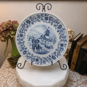 May include: A round white ceramic plate featuring a blue transferware scene of a winter landscape with a windmill, a frozen canal, and figures ice skating. The border is decorated with a detailed pattern of blue flowers and scrolling foliage.