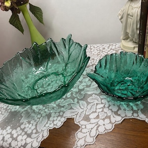 Op de afbeelding: Twee smaragdgroene glazen schalen in de vorm van bladeren met een getextureerd oppervlak en geschulpte randen. De grotere schaal is langwerpig en ovaal, terwijl de kleinere schaal ronder is. Beide stukken hebben een doorschijnende afwerking.