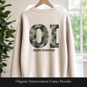 May include: A beige crewneck sweatshirt featuring a large, bold graphic on the back. The graphic consists of the letters OI filled with a green, brown, and black camouflage pattern, with the text ORGANIC INTERVENTION printed in dark green below.