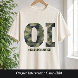 May include: A cream-colored short-sleeve t-shirt featuring a large graphic of the letters OI filled with a green, brown, and black camouflage pattern. Below the letters, the text ORGANIC INTERVENTION is printed in a dark green sans-serif font.
